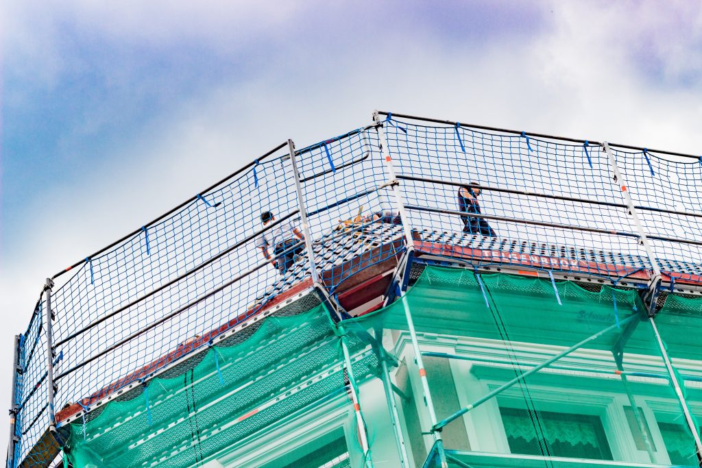 Dachdecker bei der Arbeit mit einem gut abgesicherten Gerüst im Vordergrund